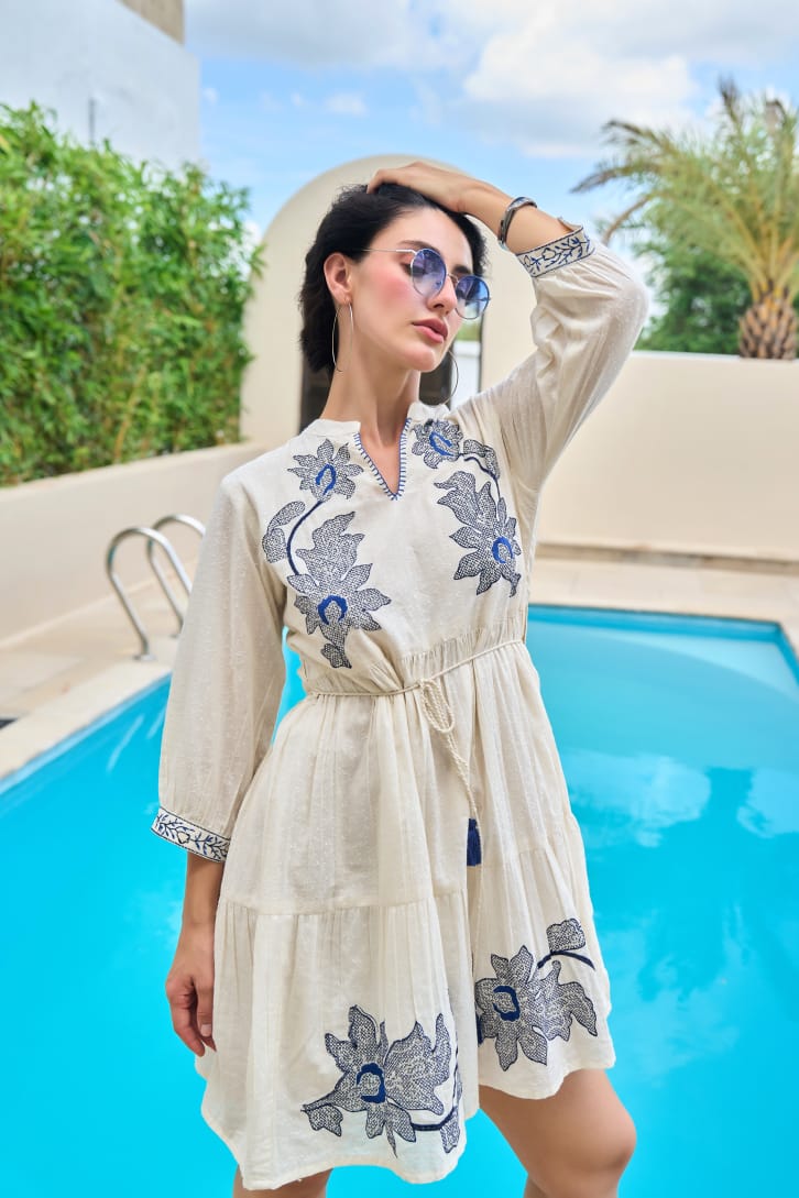 Woman wearing an ivory short boho-style dress with blue floral embroidery, V-notch neckline, drawstring waist, and 3/4 sleeves, posing by a pool.